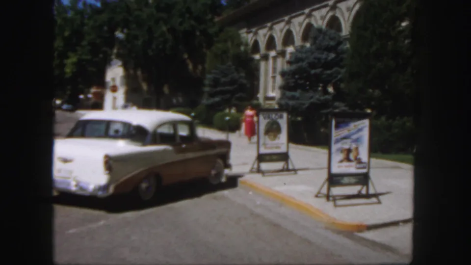 1958 Colorado campus exterior with a woman in a red dress, A-frame movie posters, trees, and a parked two-tone 1950s sedan.