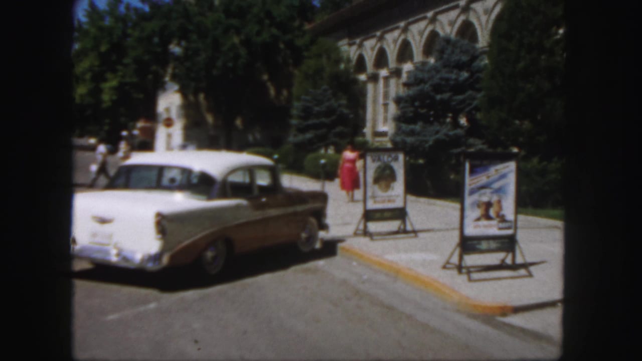 Woman in red walks along the curb near two poster stands and a parked 1950s car.