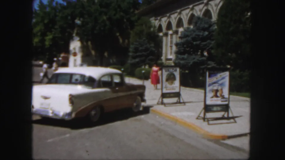 Colorado college sidewalk scene from 1958 showing a red-dressed woman near movie-poster stands and a parked white-and-brown sedan.