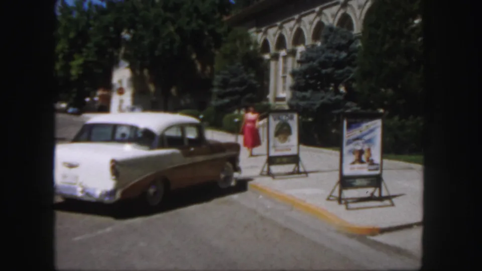 1958 Colorado campus view with a woman in red near A-frame poster signs, a two-tone Chevrolet-style sedan, trees, and arched windows.