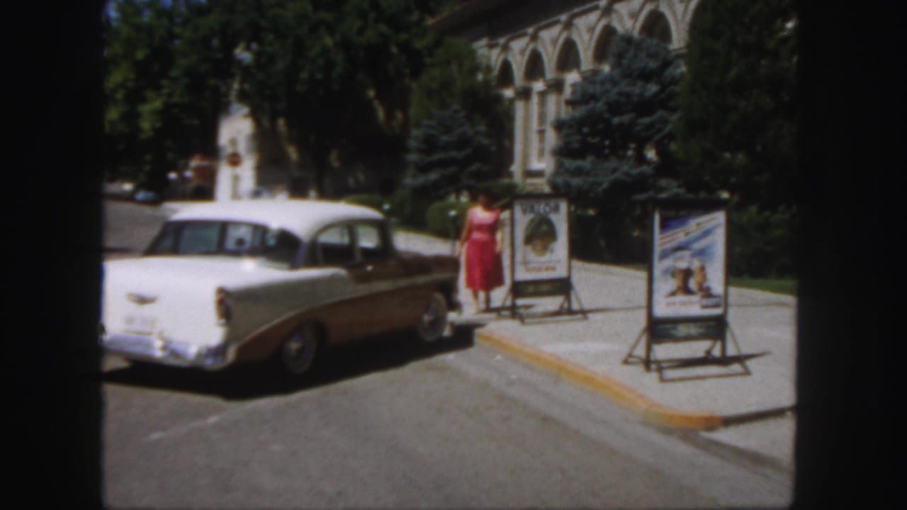 Woman in a red dress stands beside a two-tone sedan near sidewalk movie posters.