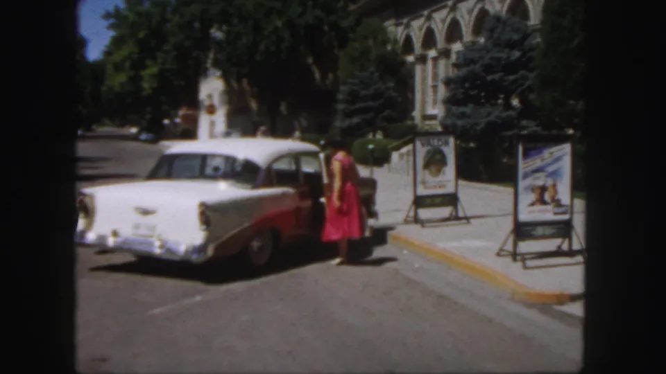 1958 Colorado frame showing the woman in red at the sedan door, with roadside posters and a large arched facade behind her.