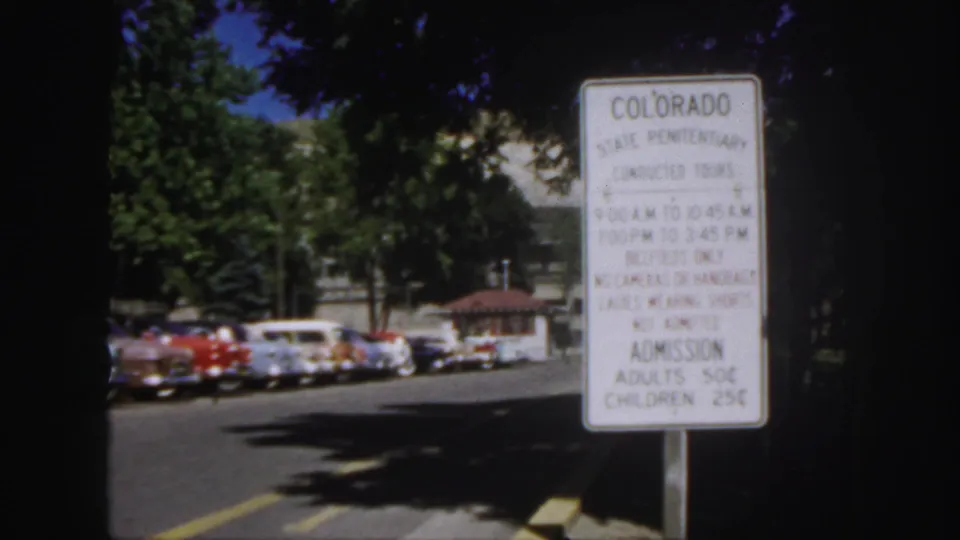 1958 Colorado frame showing a State Penitentiary tour-hours and admission sign, with trees and parked mid-century cars beyond it.