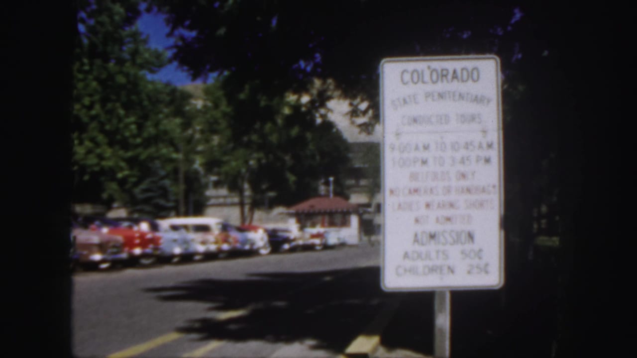 Colorado State Penitentiary tour sign beside a road with parked 1950s cars and trees.