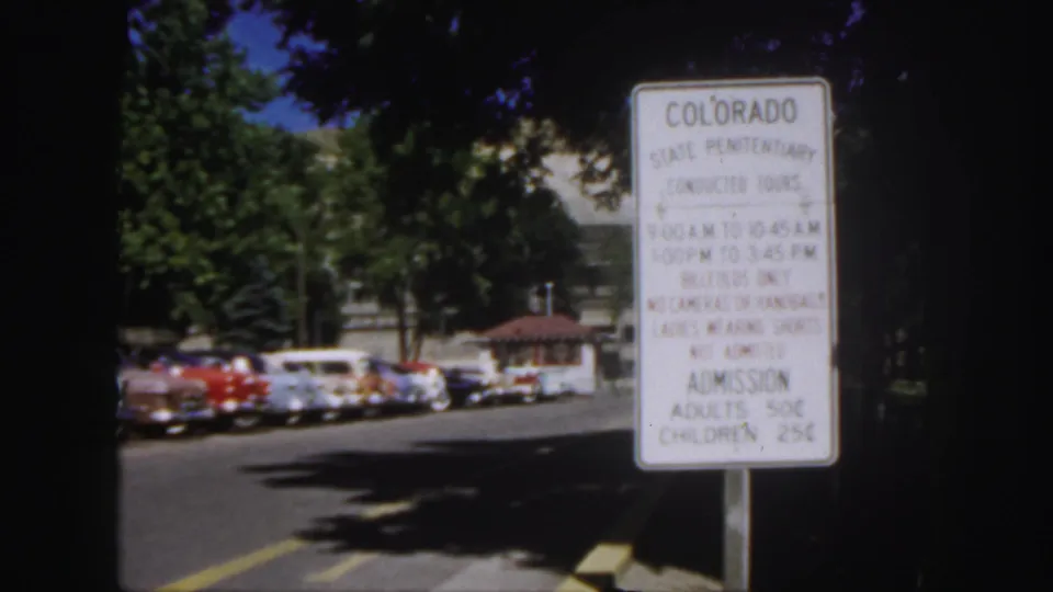 1958 Colorado frame with the penitentiary conducted tours sign in the foreground and 1950s cars parked under trees.