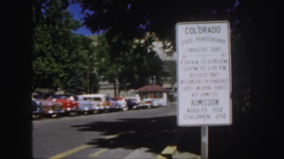 Color 8mm still of the Colorado State Penitentiary tour sign, with parked 1950s cars and foliage visible behind it.