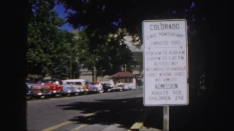 1958 Colorado home-movie frame holding on a penitentiary tour sign, with mid-century cars and trees behind it.