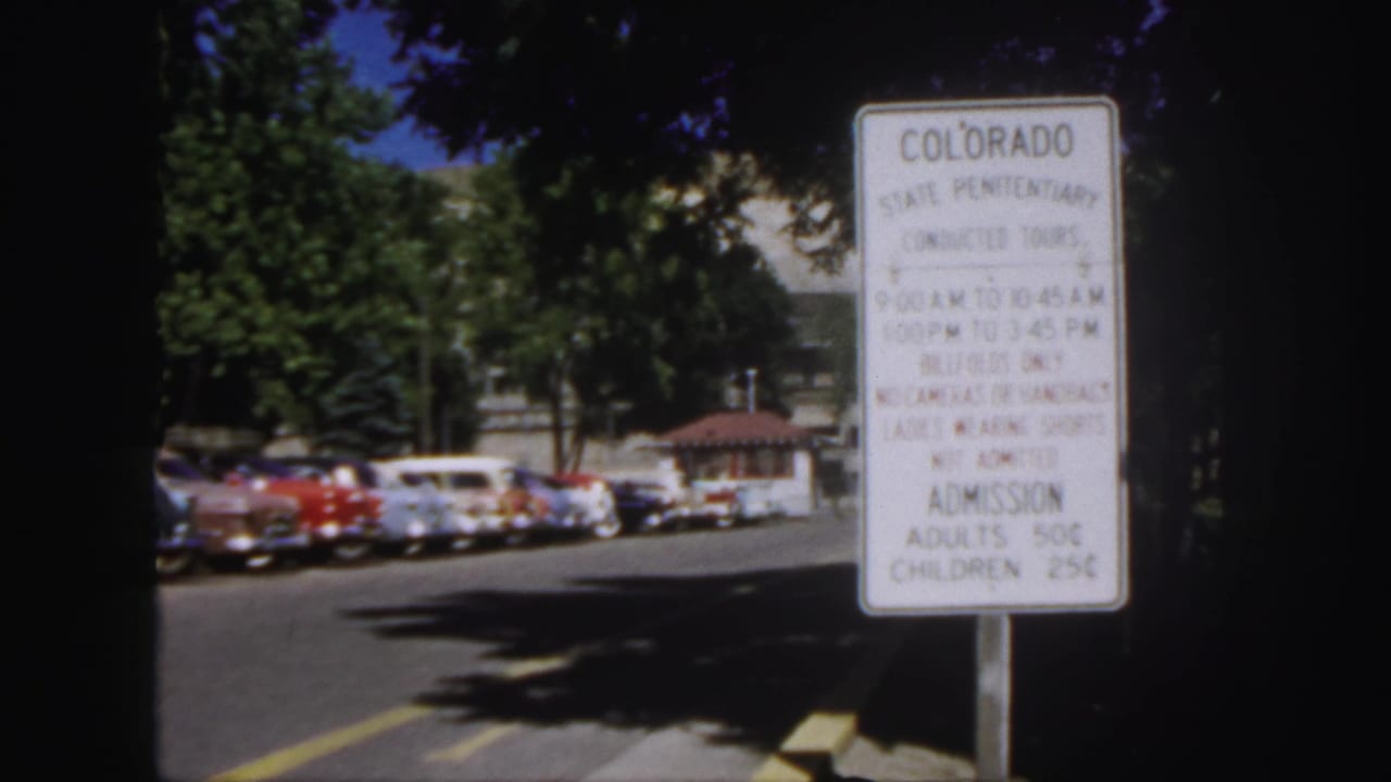 Colorado State Penitentiary tour sign beside a road with parked 1950s cars and trees.