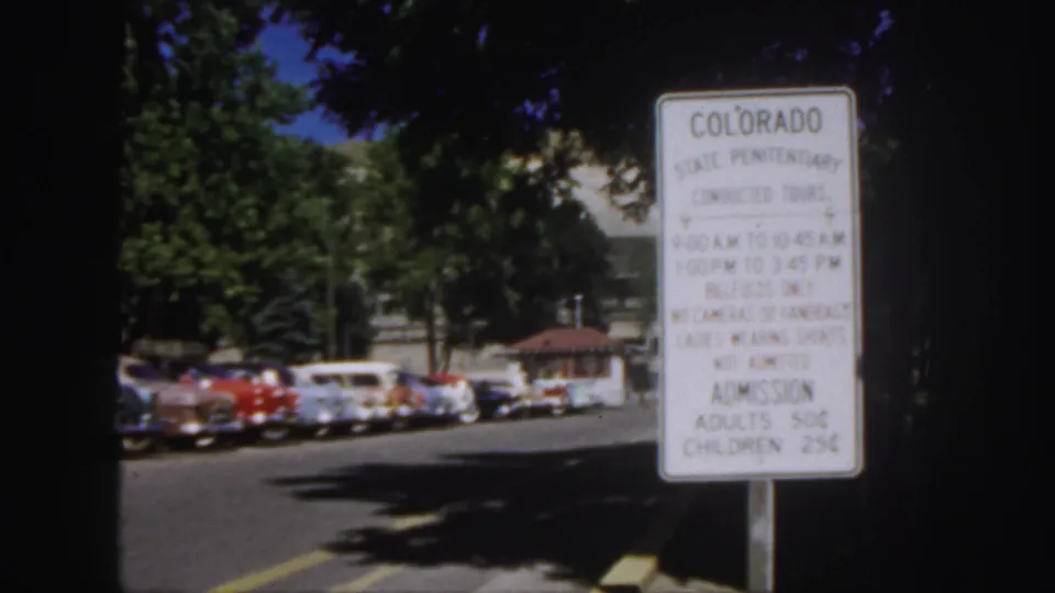 Colorado State Penitentiary sign remains in view, with 1950s cars parked along the road and trees shading the institutional grounds.