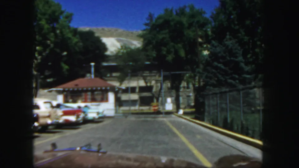 Visitor-shot view toward the Colorado State Penitentiary entrance area, with booth, fencing, roadway, parked car, trees, and hillside beyond.