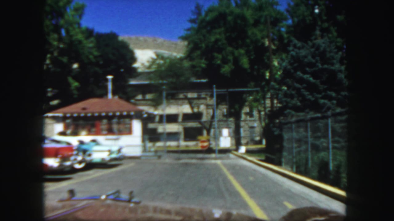 Car-window view of a resort checkpoint with a red-roofed booth, parked cars, fence, trees, and mountain behind.