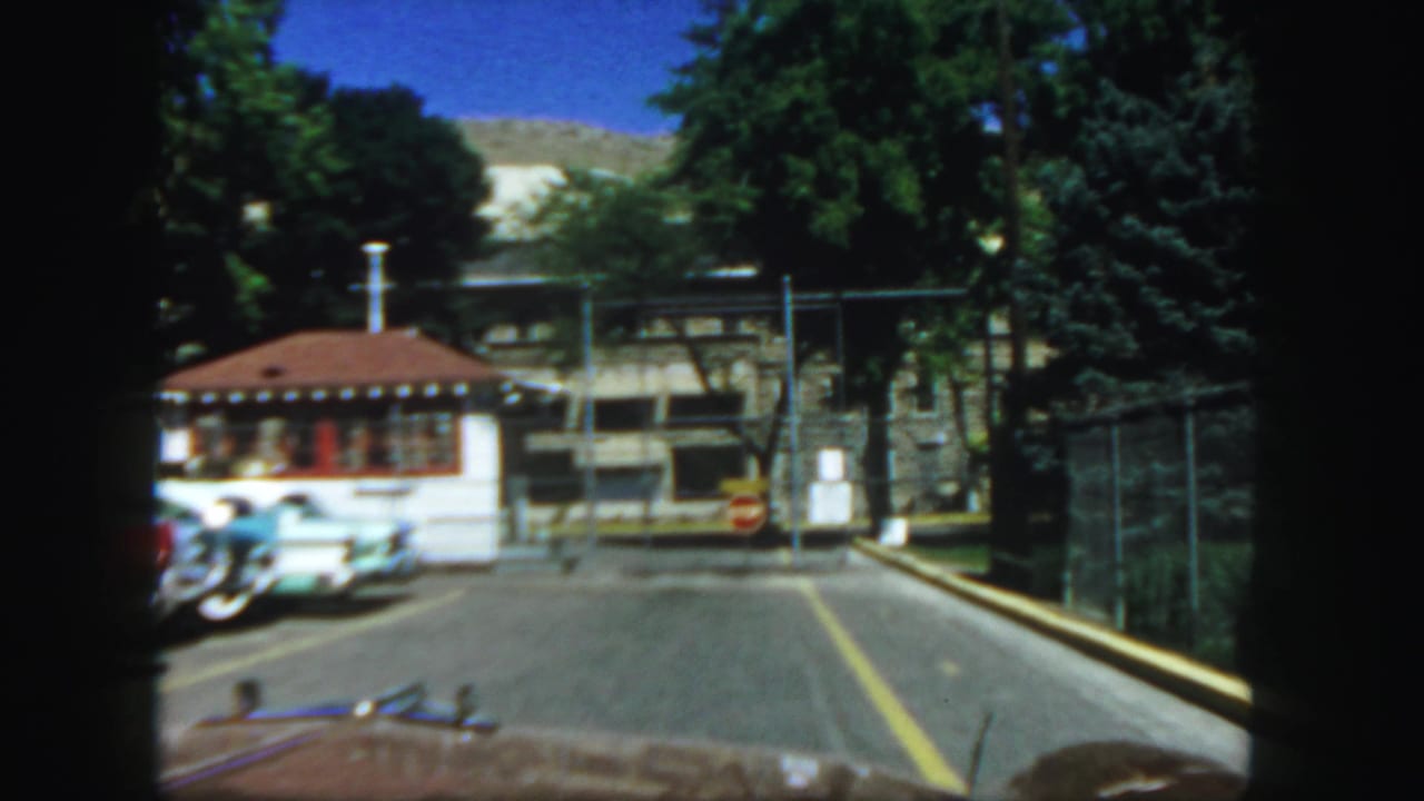 Roadway beside a small booth and parked cars, with fence, stop sign, trees, and mountain in the background.