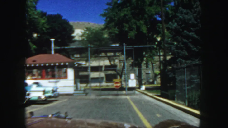 1958 Colorado home-movie frame gives a closer view of a red-roofed checkpoint booth, fenced grounds, stop sign, and road from a car.