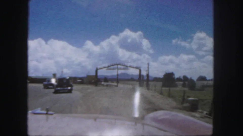 Colorado 8mm travel frame shows an open road from inside a car, another vehicle ahead, roadside fencing, open land, and dramatic clouds.