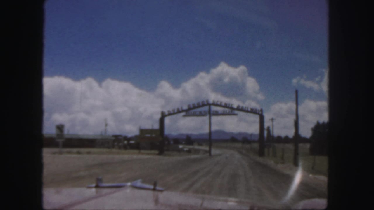 Dirt road bends toward a large arched sign with clouds and mountains beyond.