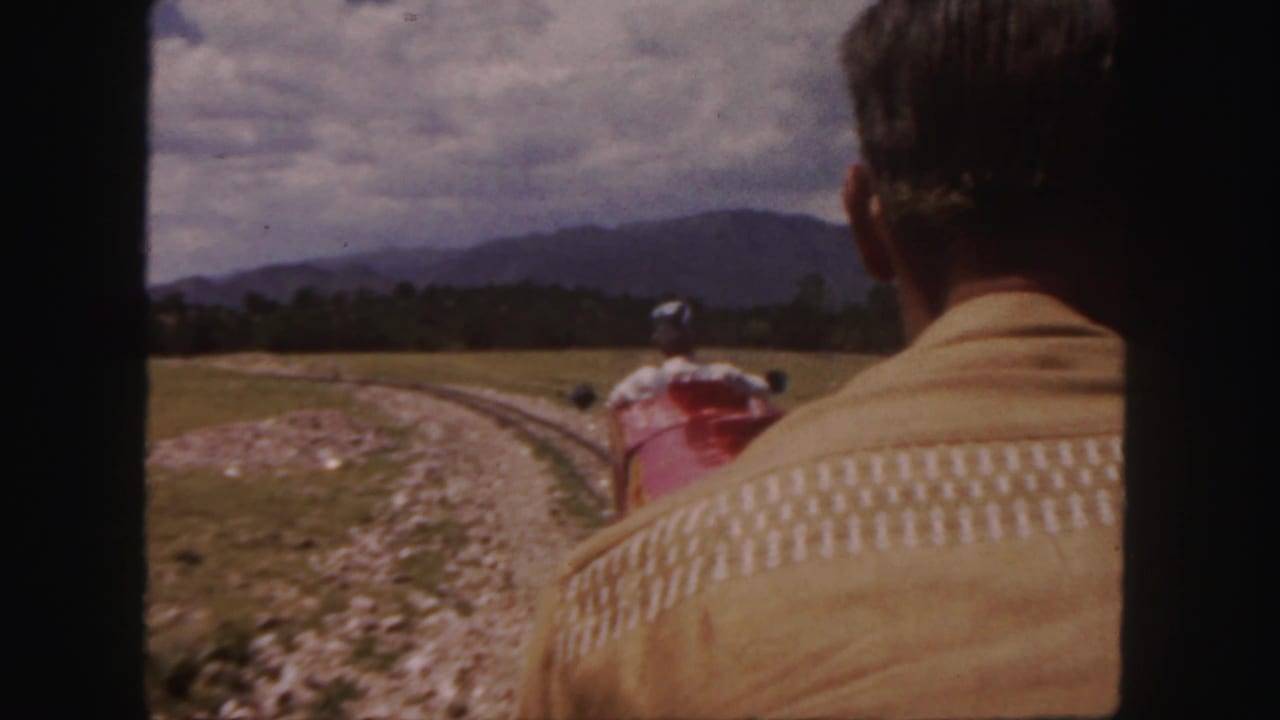 1958 Colorado home-movie view from a moving train, with a seated man in front and a red train car ahead on a curved rural track.