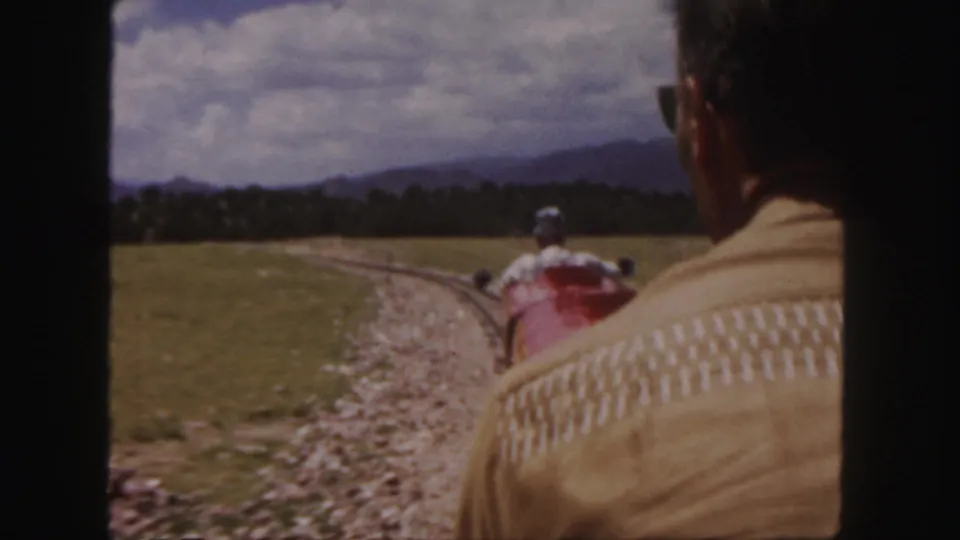 Home-movie frame from a moving 8mm train ride, looking past a seated passenger toward a red car and rough track in Colorado scenery.