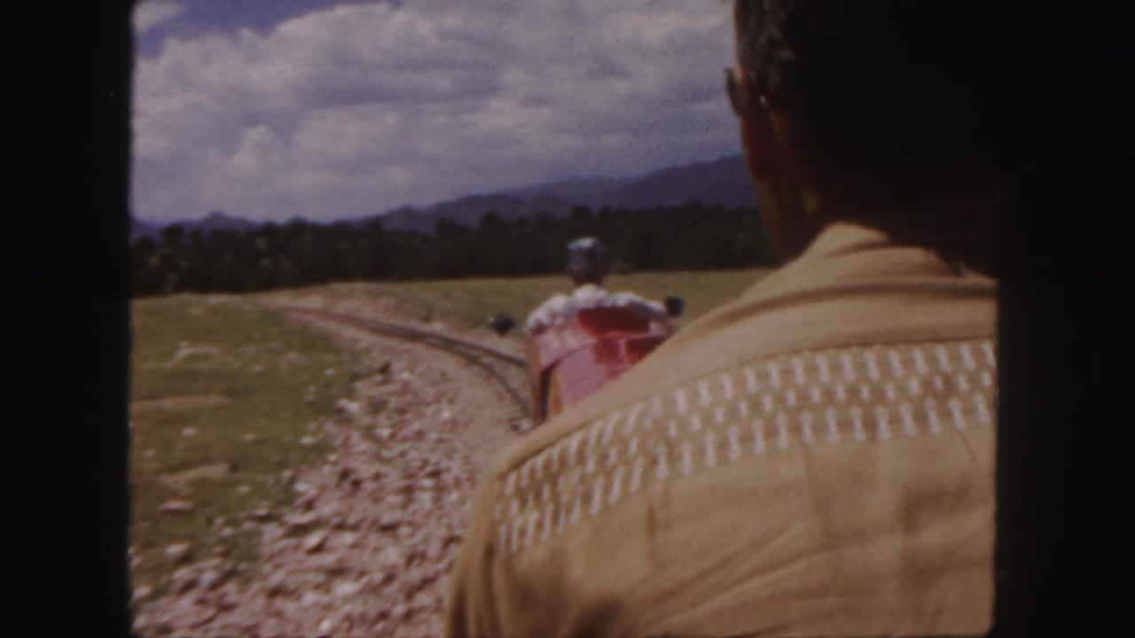 In 1958 Colorado 8mm footage, an adult in a beige patterned jacket faces a red miniature train moving through open grassland toward distant mountains.
