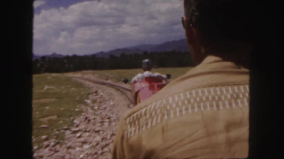 In 1958 Colorado 8mm footage, an adult in a beige patterned jacket faces a red miniature train moving through open grassland toward distant mountains.