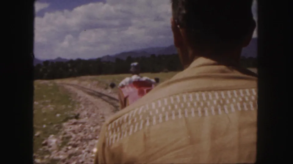 The Colorado home-movie frame shows a red miniature train ahead of a back-facing adult passenger, with rails curving through grass below a partly cloudy sky.