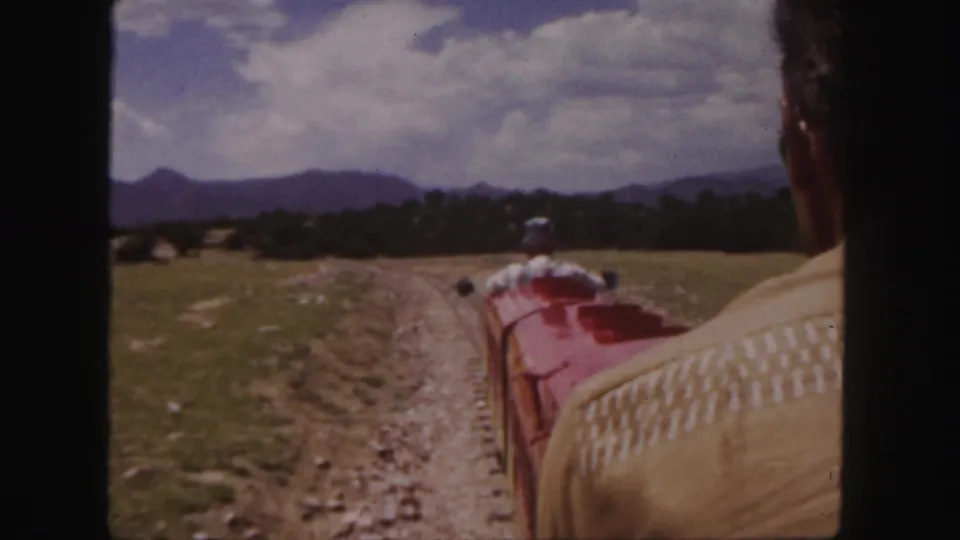 Near the end of the clip, the red miniature train curves away from the camera beside a back-facing adult passenger in the Colorado landscape.