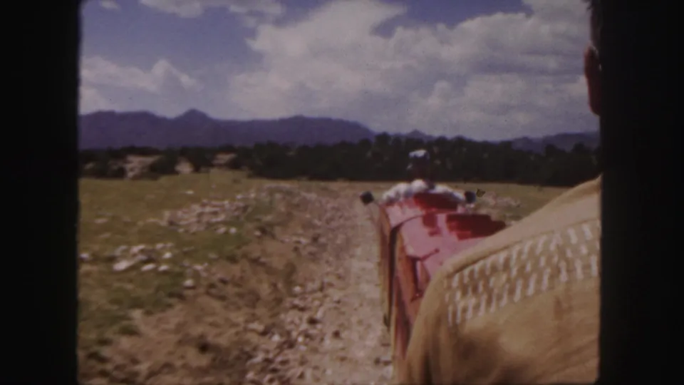 Rider's-eye 1958 Colorado view of a red miniature train traveling along a rocky dirt path with trees and mountains beyond.