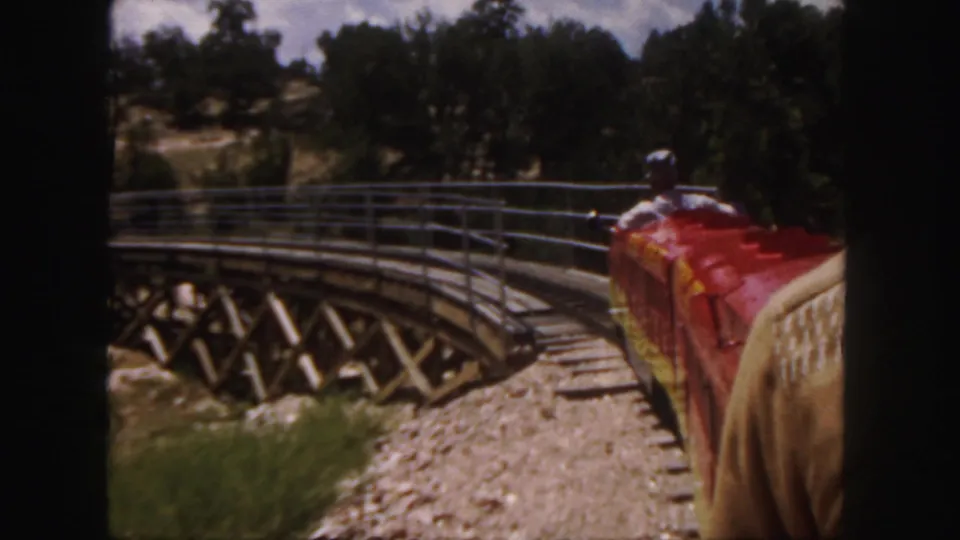 1958 Colorado home-movie frame showing the red miniature train nearing a curved wooden trestle bridge in a wooded area.