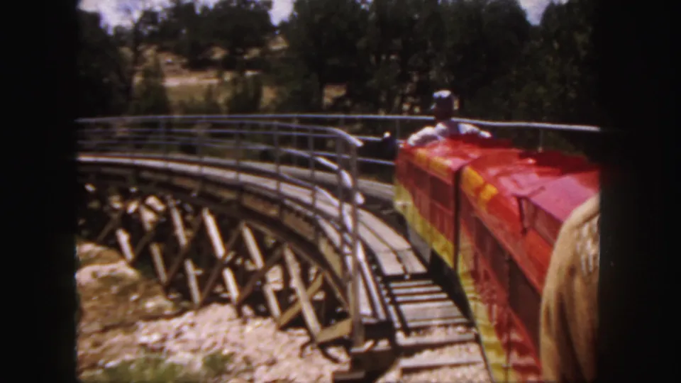 Rider's-eye 1958 Colorado 8mm view of a red miniature train crossing a curved wooden trestle bridge surrounded by trees.