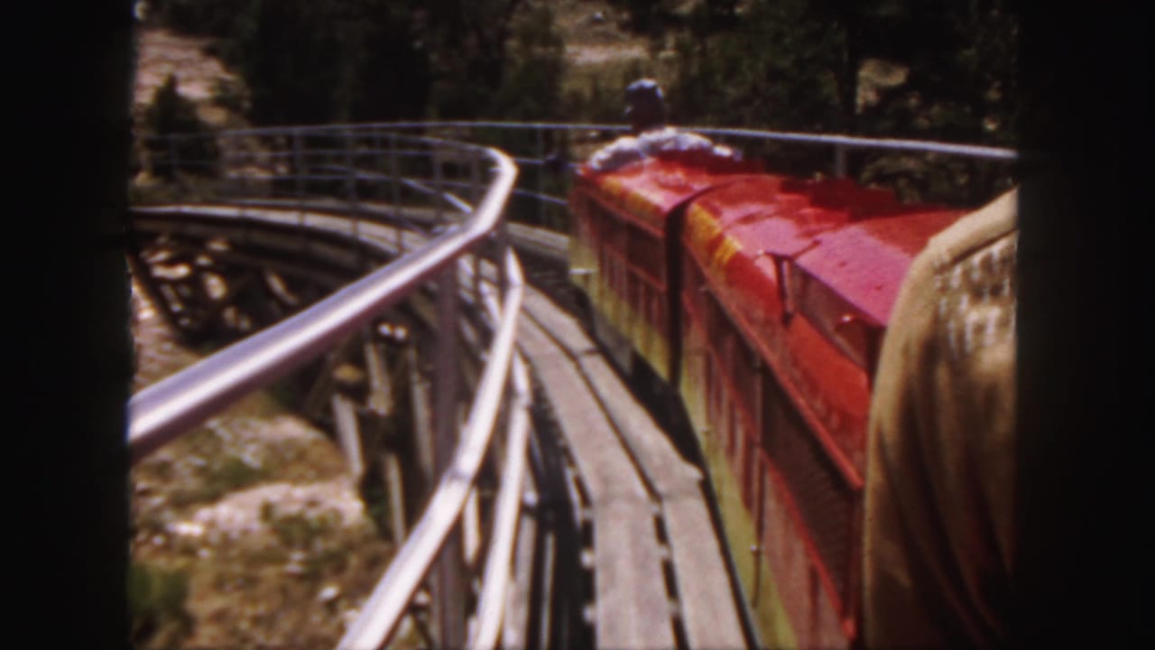 Passenger view of a red miniature train on a curve, with railings, trees, and an adult rider visible.