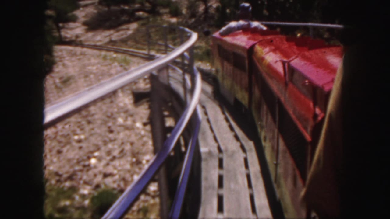 1958 Colorado home-movie frame of a red open-air train car on a curved track through dry rocky ground.