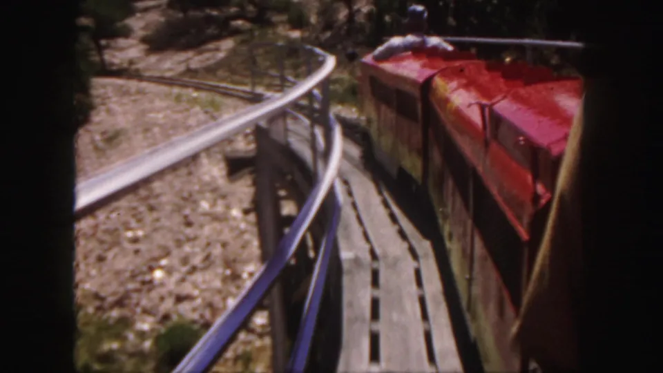1958 Colorado home-movie frame of a red open-air train car on a curved track through dry rocky ground.