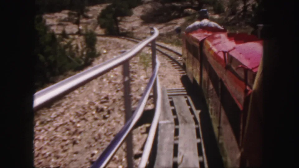 Red open-air train car on a curve in Colorado terrain, seen from a moving 8mm home-movie viewpoint.