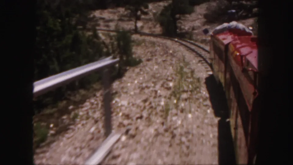 Moving 8mm view from near the train, showing curved track, metal rail, rocky ground, and the red car edge.