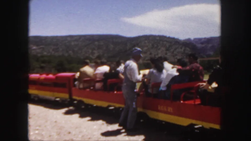 1958 Colorado tourist train scene with seated passengers, a standing uniformed man, red cars, and distant hills.