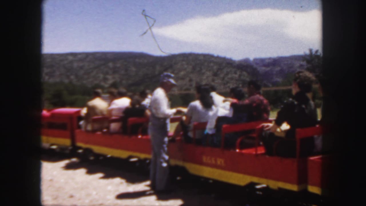 1958 Colorado home-movie frame showing passengers on an open red and yellow tourist train with a standing man in a cap nearby.