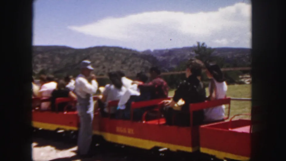 1958 Colorado frame of an open tourist train paused or moving slowly with visitors seated and a capped man beside the car.