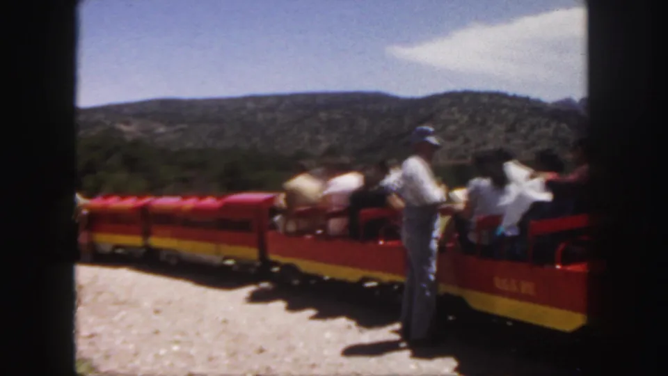 1958 Colorado home-movie frame showing the open tourist train from the side as it stretches across a sunny scenic setting.