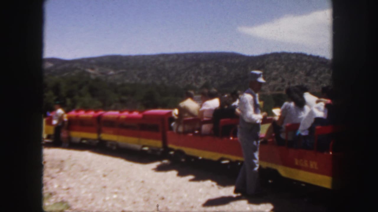 1958 Colorado 8mm frame showing a red and yellow open-air tour train, seated visitors, and a man in a blue shirt and cap beside the cars.