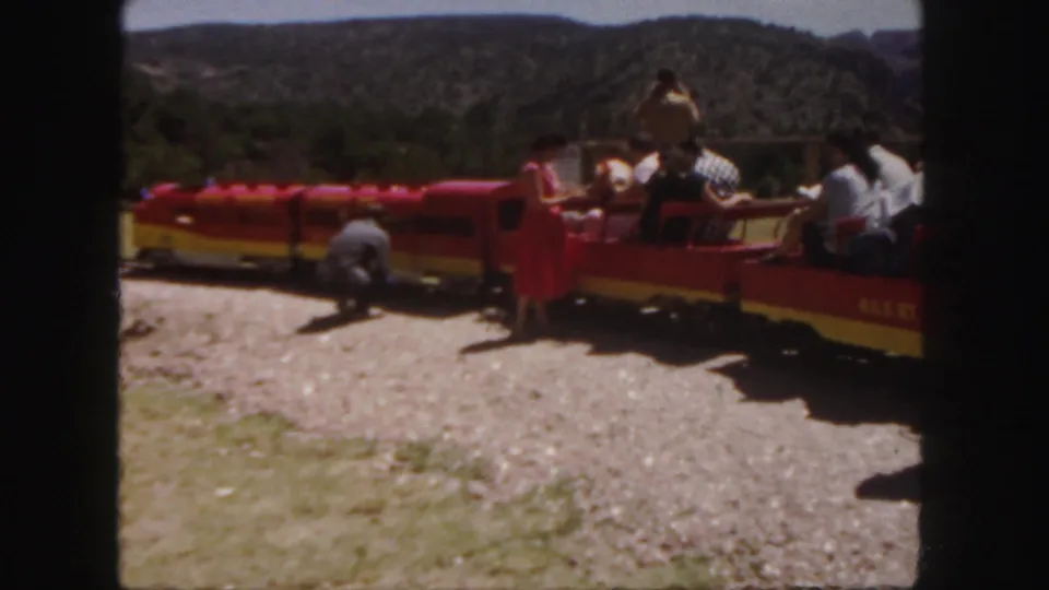 Open-air red and yellow tour train in Colorado with passengers seated in the cars and a person bending near the front on a sunny day.