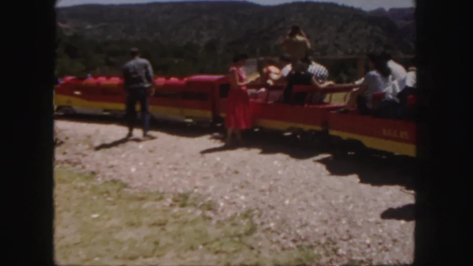 A man stands near a red and yellow open-air tour train while passengers sit in the cars, with Colorado hills visible behind them.