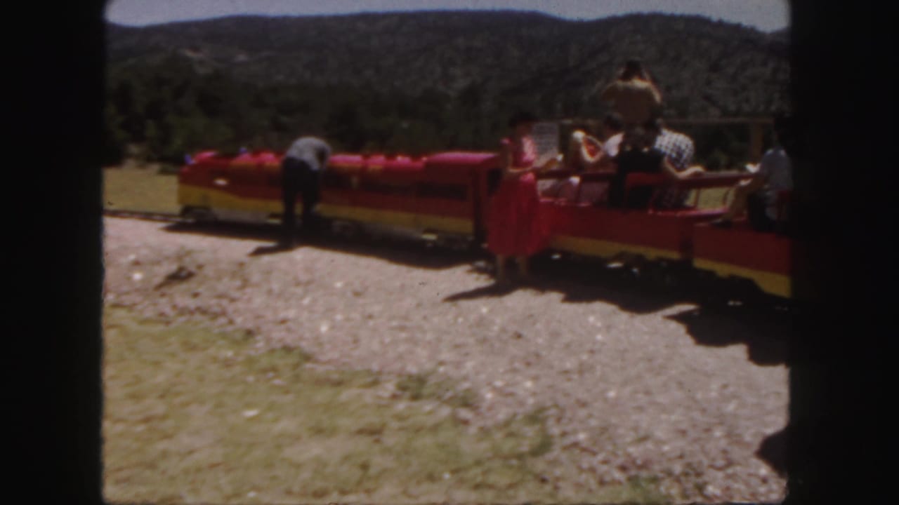 People gathered around a red and yellow miniature train on a gravel track.