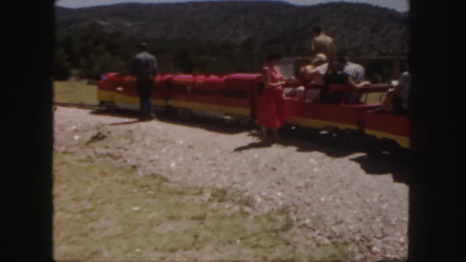 Passengers sit in open red train cars at a Colorado outdoor attraction, with trees and hills beyond.