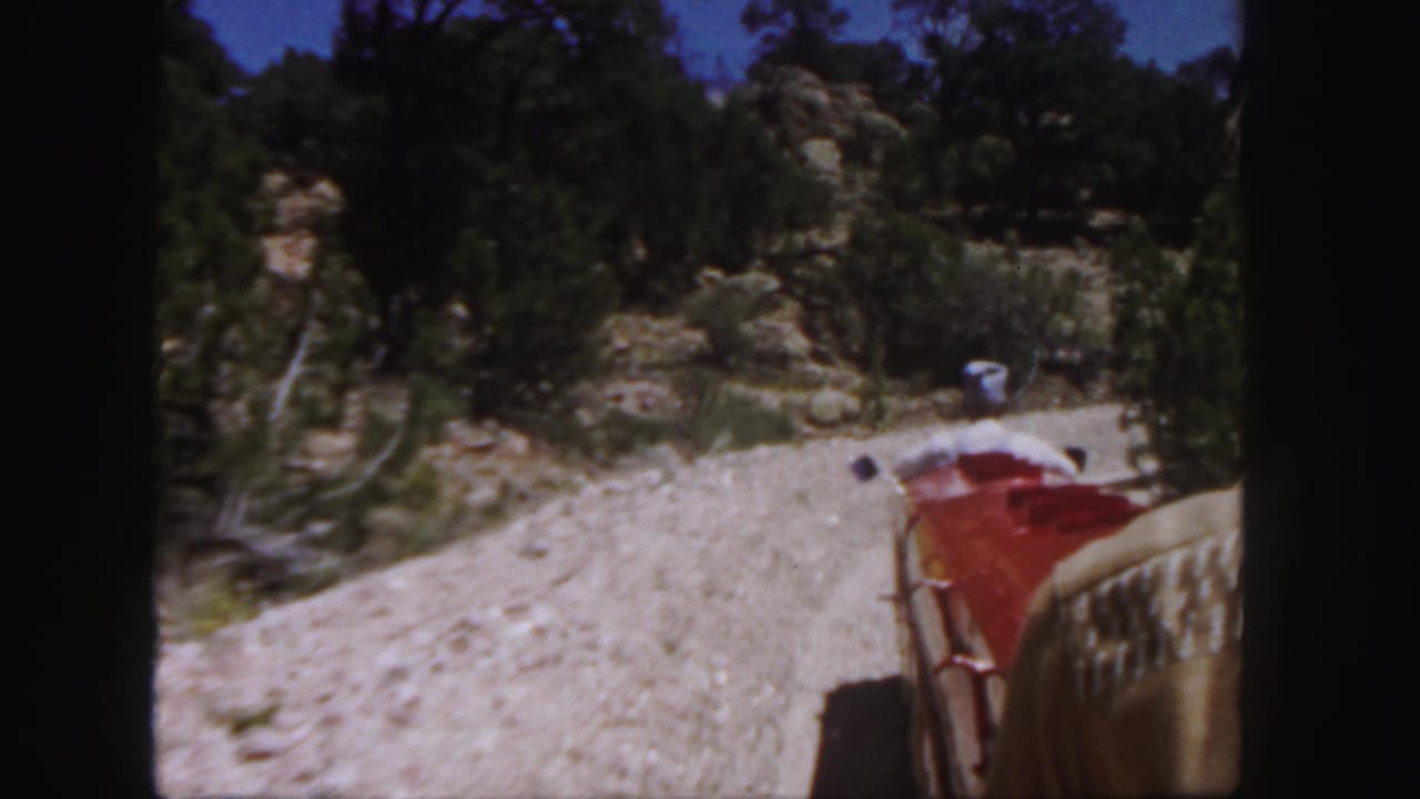 View from a red miniature train car moving along a gravel track through rocky trees.