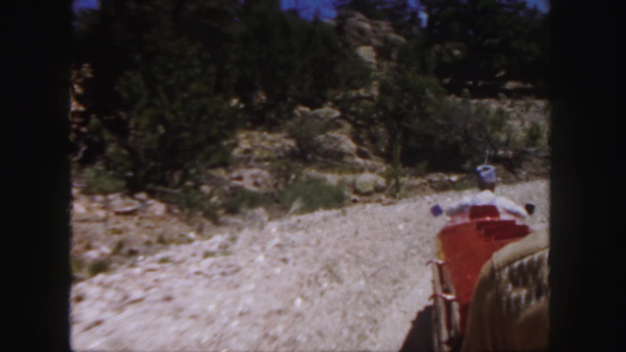 Red train car and conductor ahead on a gravel track beside rocks and trees.