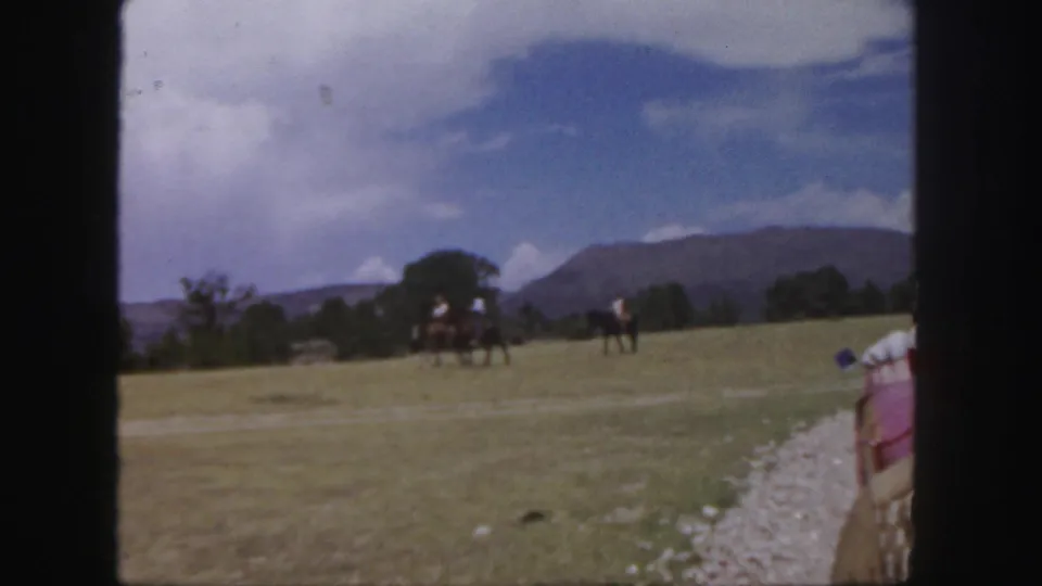Horseback riders move across an open Colorado field under clouds, with distant mountains beyond the train route.