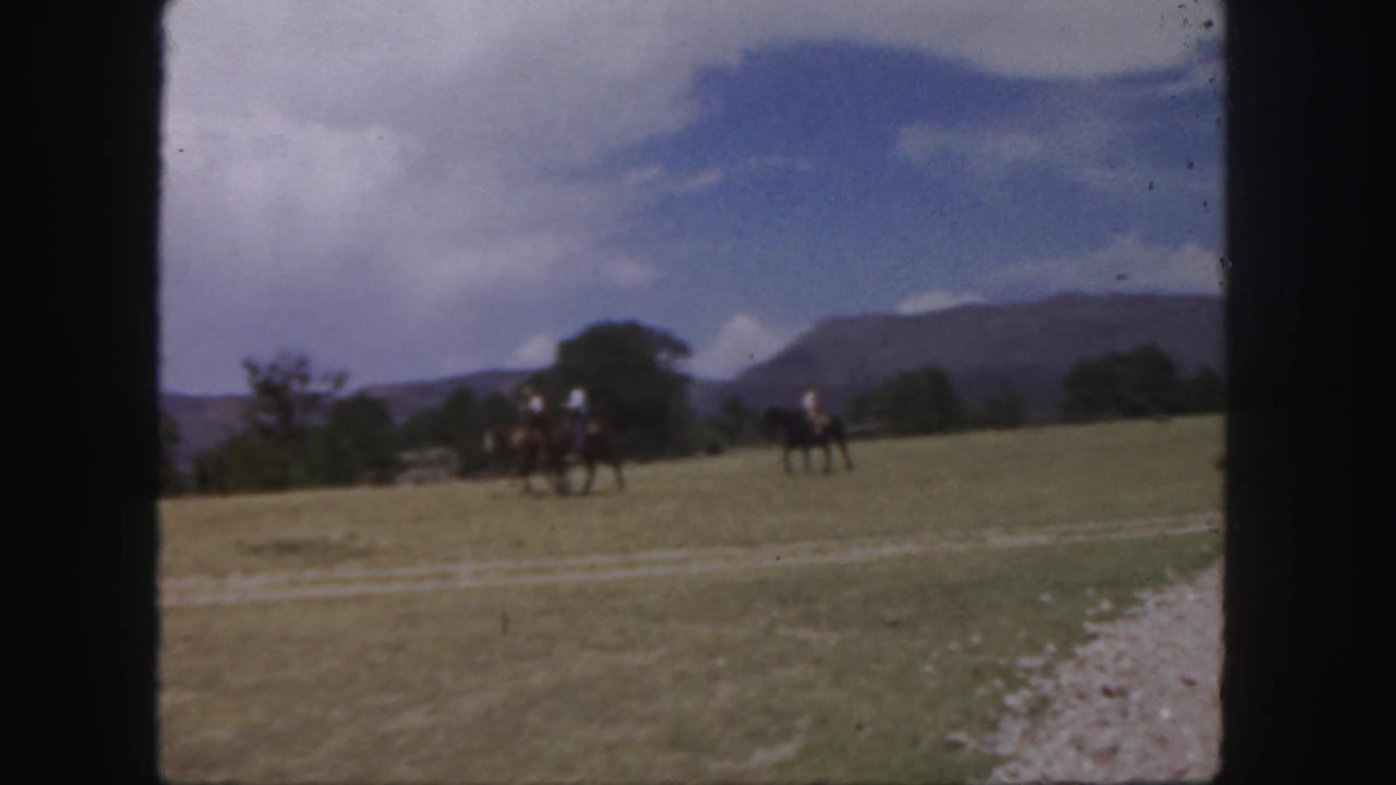 Two horseback riders cross a grassy field with trees and mountains in the distance.