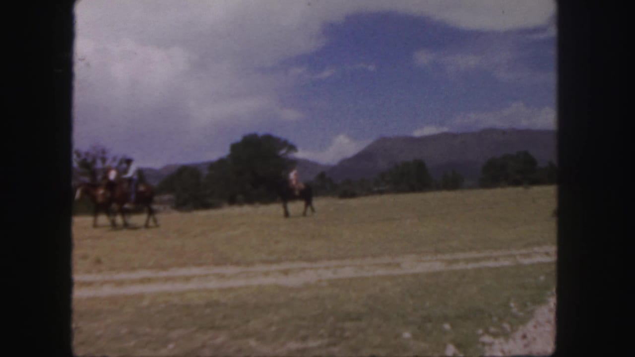 Mounted riders continue across a field, framed by trees, mountains, and a clouded sky.