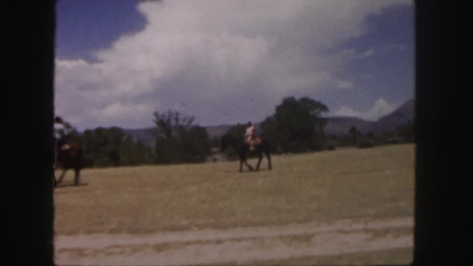 A soft 8mm home-movie frame shows horseback riders spread across a Colorado field with trees, mountains, and large clouds behind them.