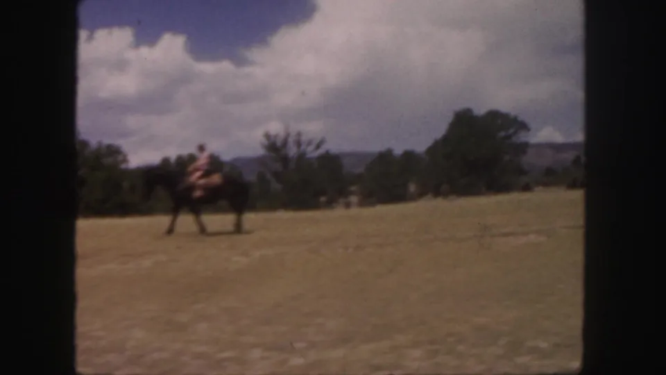 A rider on horseback crosses the foreground of a Colorado field while another mounted figure is partly visible near the frame edge.
