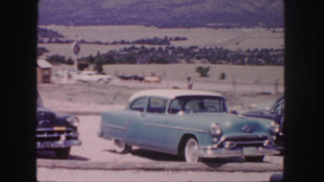 Classic cars parked beside a rural road with open fields and distant hills in the background.
